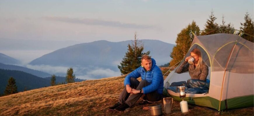 woman hiker sitting inside camp tent and drinking coffee while man looking at tourist gas burner couple travelers resting on grassy hill and drinking hot beverage while hiking together in mountains
